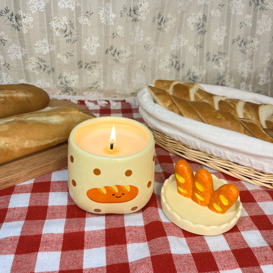 Lit baguette & butter scented candle in a ceramic jar with polka dots and a smiling baguette illustration, styled on a red gingham tablecloth with its baguette-shaped lid, fresh bread loaves, and a woven basket in the background.
