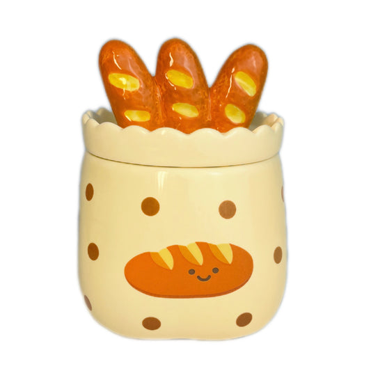 Baguette & butter scented candle in a ceramic jar with polka dots and a smiling baguette illustration, topped with a baguette-shaped lid, photographed on a white background.