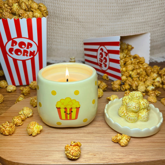 Lit caramel popcorn scented candle in a ceramic jar with yellow polka dots and a smiling popcorn bucket illustration, styled on a wooden surface with scattered popcorn, a popcorn-shaped lid, and red-and-white popcorn boxes in the background.