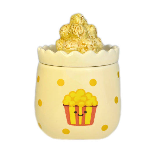 Caramel popcorn scented candle in a ceramic jar with yellow polka dots and a smiling popcorn bucket illustration, topped with a popcorn-shaped lid, photographed on a white background.