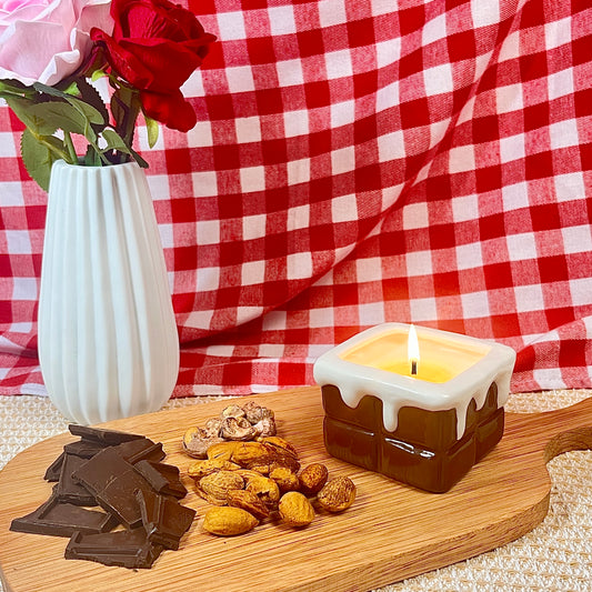 Chocolate ganache-scented ceramic soy candle with glossy square vessel, styled on a wooden board with dark chocolate and almonds against a red gingham backdrop.