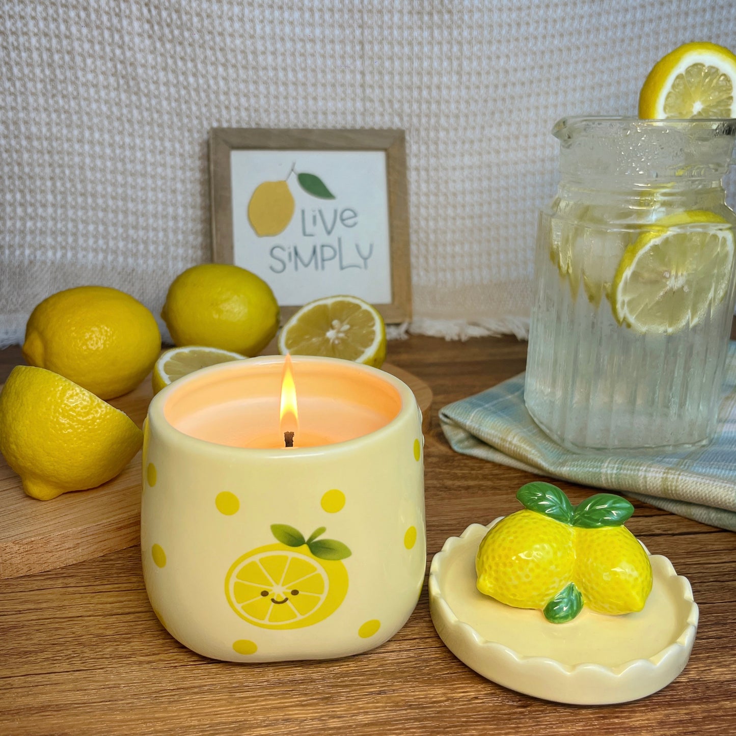 Lemon-themed candle on a wooden surface with lemons and a glass of lemonade in the background.