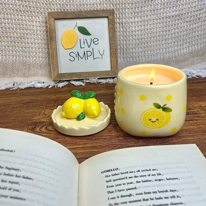 Lit sugared lemon zest scented candle in a cheerful ceramic jar with yellow polka dots and a smiling lemon illustration, styled on a wooden surface beside its lemon-shaped lid, an open book, and a framed “Live Simply” sign in the background.