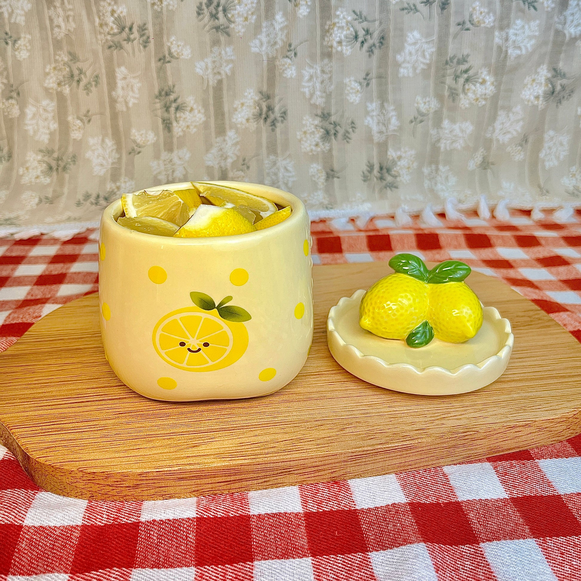 Repurposed ceramic candle jar with yellow polka dots and a smiling lemon illustration, reused as a small container holding fresh lemon slices, with the lemon-shaped lid placed beside it on a wooden board and red gingham tablecloth.