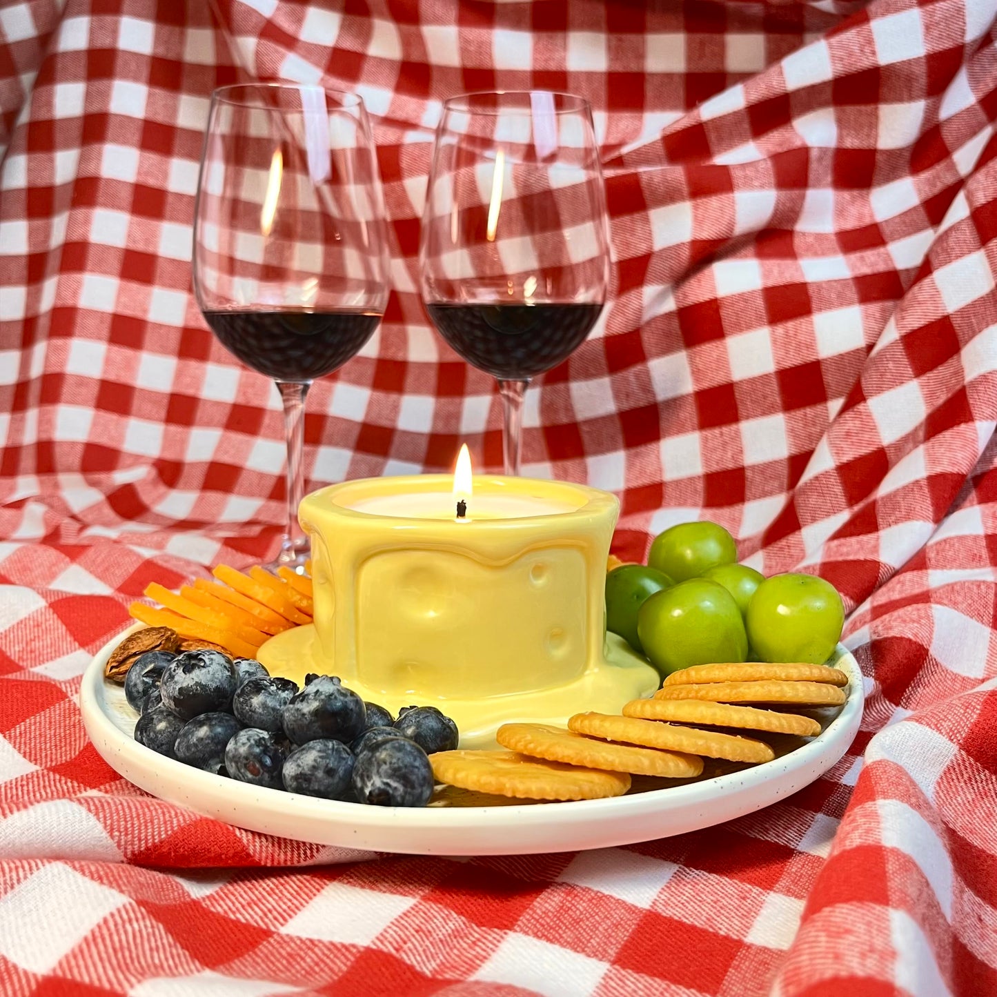 Vanilla cheesecake-scented ceramic soy candle styled like a cheese wheel on a plate with crackers, grapes, blueberries, and wine glasses.