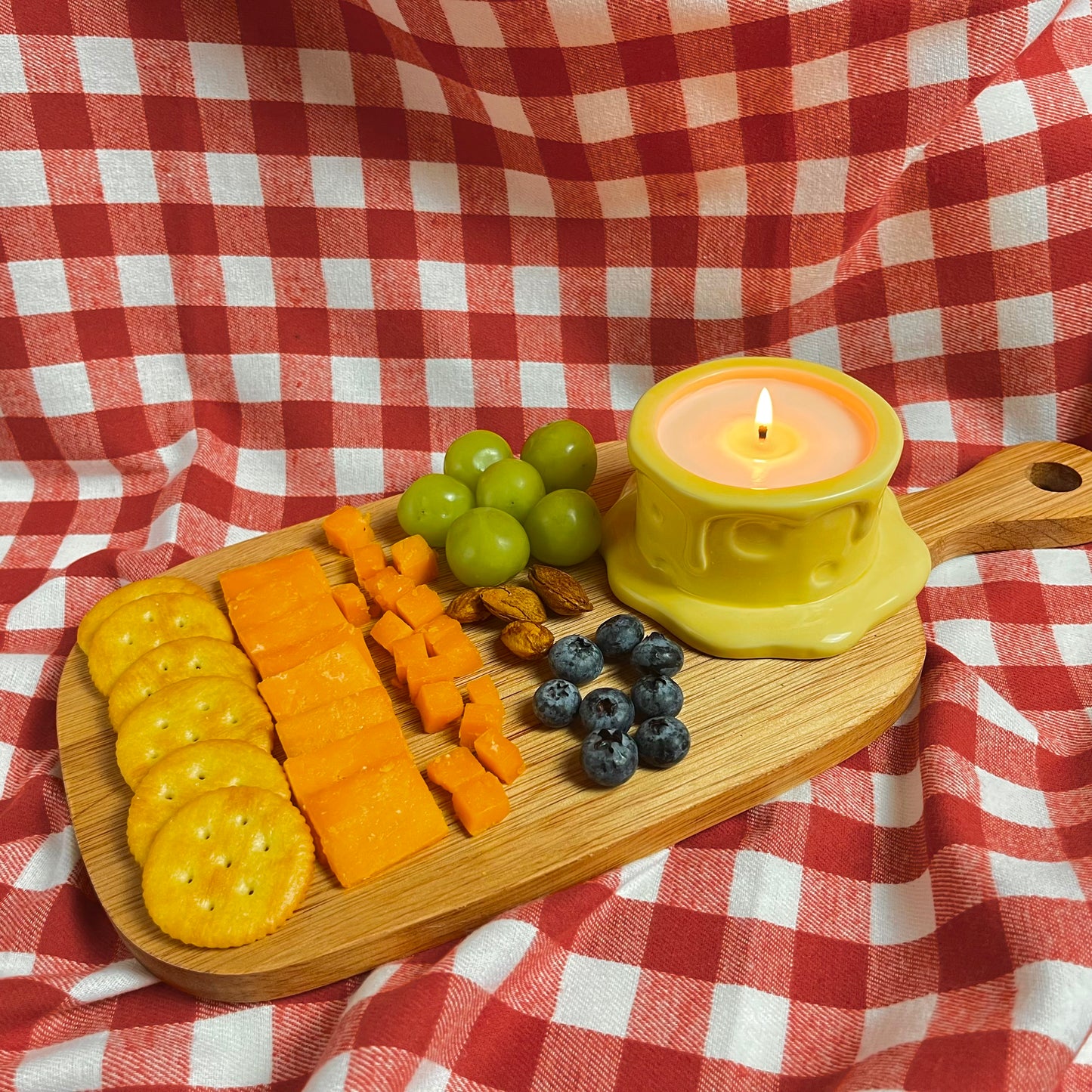 Vanilla cheesecake-scented ceramic soy candle styled on a wooden cheese board with crackers, cheese, grapes, and berries on a red gingham cloth.