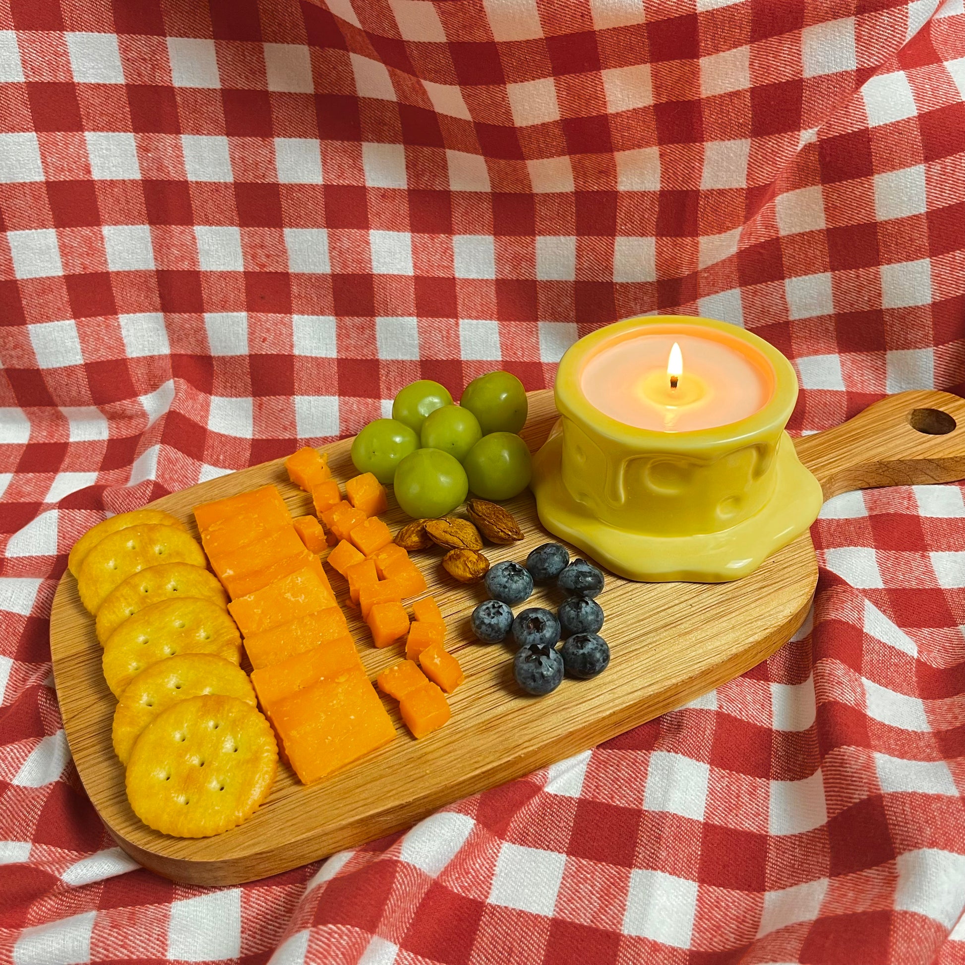 Vanilla cheesecake-scented ceramic soy candle styled on a wooden cheese board with crackers, cheese, grapes, and berries on a red gingham cloth.
