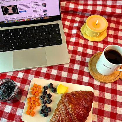 Vanilla cheesecake-scented ceramic soy candle styled on a red gingham tablecloth beside a laptop, coffee, and breakfast pastries for a cozy work-from-home scene.