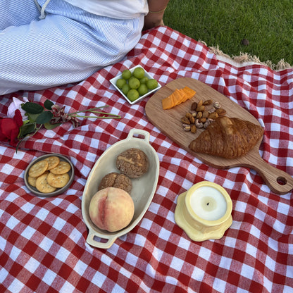 Vanilla cheesecake-scented ceramic soy candle styled on a red gingham picnic blanket with pastries, fruit, cheese, and snacks for a cozy outdoor picnic scene.