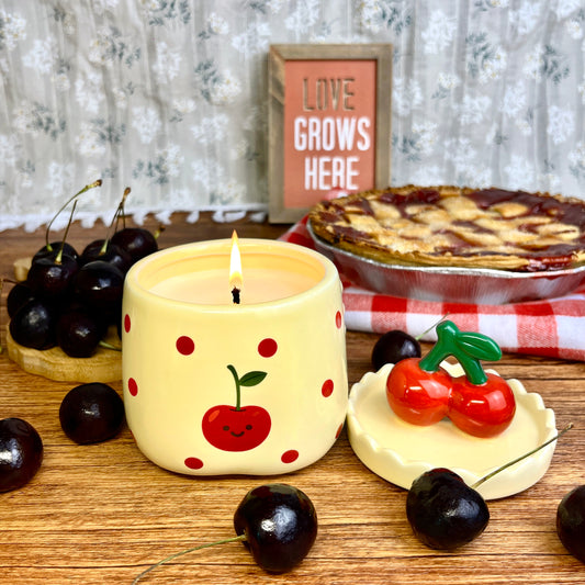 Lit velvet cherry scented candle in a ceramic jar with red polka dots and a smiling cherry illustration, styled on a wooden surface with its cherry-shaped lid, fresh cherries, and a cherry pie in the background.